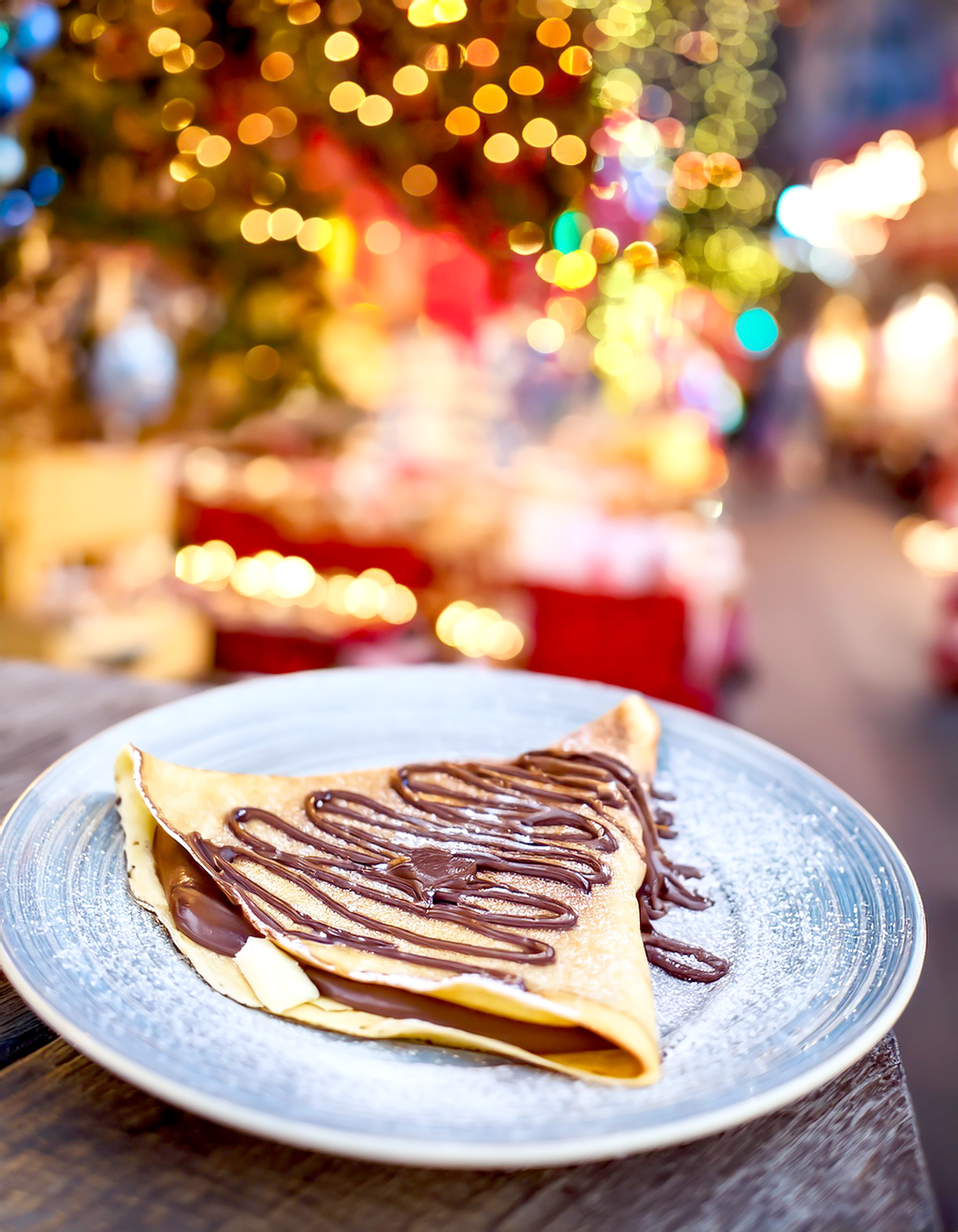 Crepes auf dem Weihnachtsmarkt Ein Crepes mit Schokosoße auf einem weißen Teller, im Hintergrund weihnachtliche Beleuchtung